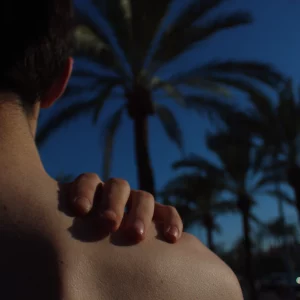 Una mano apoyada en el hombro de otra persona, con palmeras y cielo azul al fondo