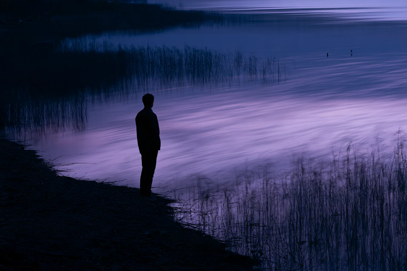 Silueta de una persona en la orilla de un lago oscuro de noche, mirando el agua bajo una luz lila fría