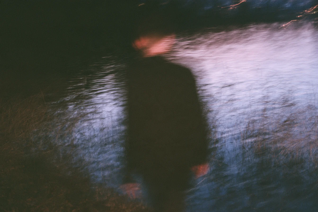 Silueta desenfocada de una persona caminando junto a un agua oscura con reflejos lila y rosa