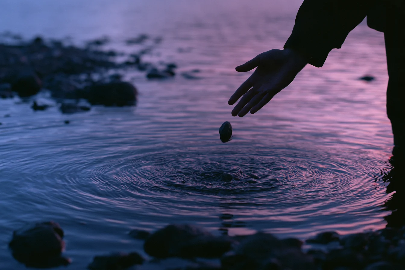 Mano de una persona dejando caer una piedra en un agua oscura con reflejos lila